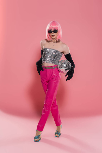 Trendy drag queen in sunglasses and shiny top posing with disco ball on pink background  - Fotografie, Obrázek