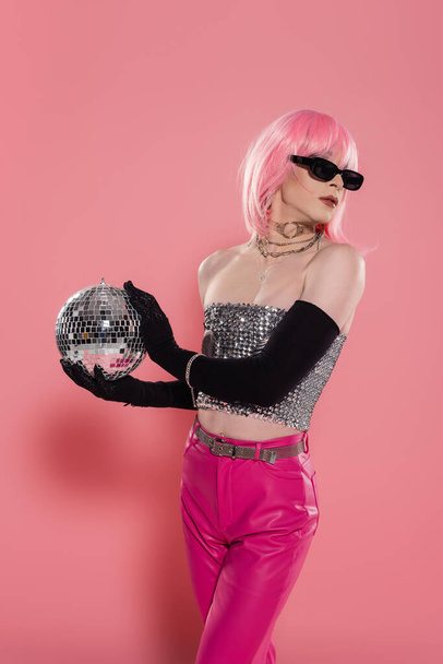 Fashionable drag queen in sunglasses and silver top holding disco ball on pink background  - Фото, изображение