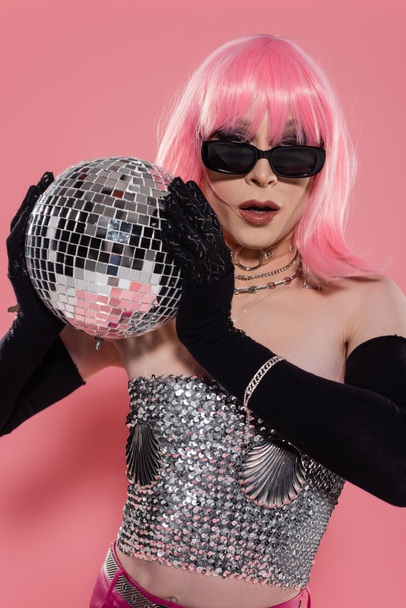 Portrait of stylish drag queen in sunglasses holding disco ball on pink background  - Φωτογραφία, εικόνα