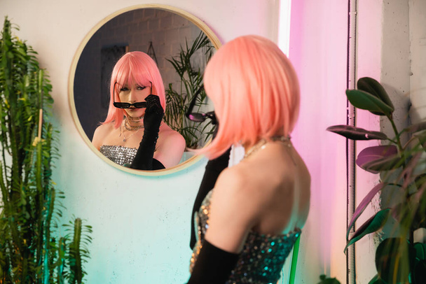 Homályos drag queen parókában napszemüveget visel a tükör közelében otthon  - Fotó, kép