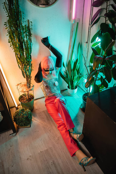 High angle view of fashionable man in gloves and shiny top sitting on floor near plants at home  - Foto, afbeelding