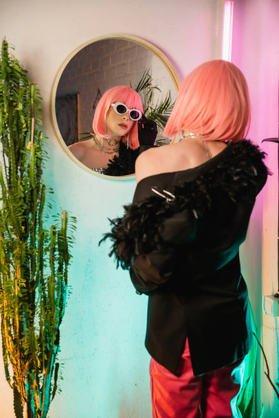 Μοντέρνο τρανσέξουαλ άτομο με σακάκι και περούκα φορώντας γυαλιά ηλίου κοντά στον καθρέφτη στο σπίτι  - Φωτογραφία, εικόνα