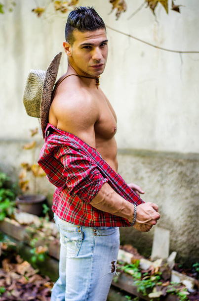 Handsome, muscular young man taking off shirt, wearing jeans and straw hat, outdoors - Foto, imagen