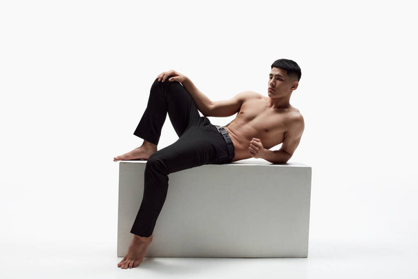 Portrait of handsome young man with muscular fit body posing shirtless in trousers against white studio background. Body-positivity. Concept of male beauty, skincare, cosmetology, mens health - 写真・画像