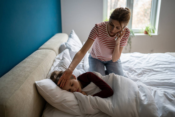 Worried young woman mother calling to pediatrician while sitting near sick son on bed. Stressed mom holding smartphone talking to doctor, feeling worried about child health, checking kids temperature - Fotografie, Obrázek