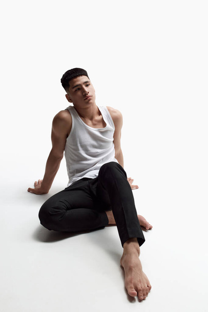 Portrait of young handsome man posing in black trousers and singlet against white studio background. Masculinity aesthetics. Concept of male beauty, skincare, cosmetology, mens health - Fotografie, Obrázek