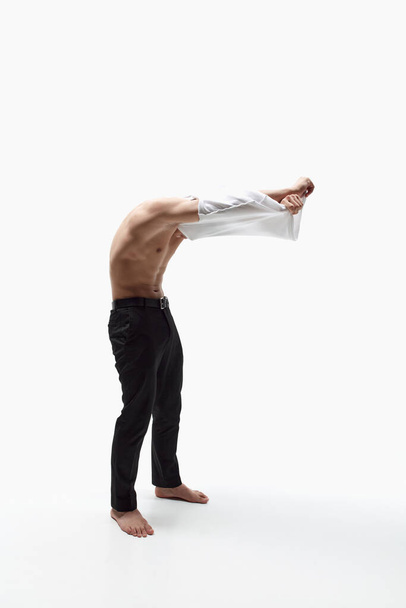 Full-length image of young muscular man taking off t-shirt, posing in trousers against white studio background. Fit, relief body shape. Concept of male beauty, skincare, cosmetology, mens health - Fotografie, Obrázek