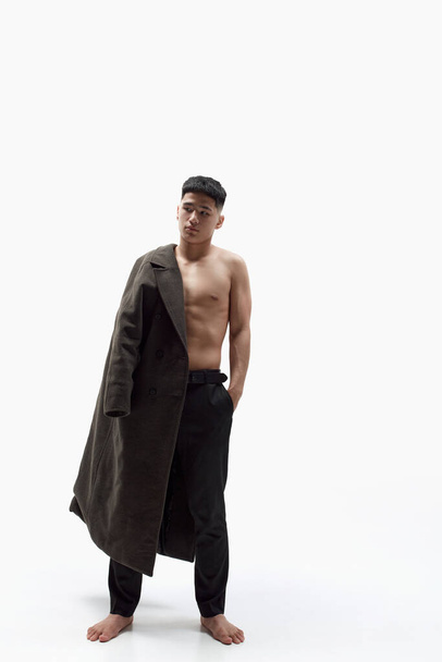 Full-length portrait of young, handsome, asian guy posing shirtless with stylish coat against white studio background. Concept of men fashion, style, body aesthetics, beauty, mens health, emotions - 写真・画像