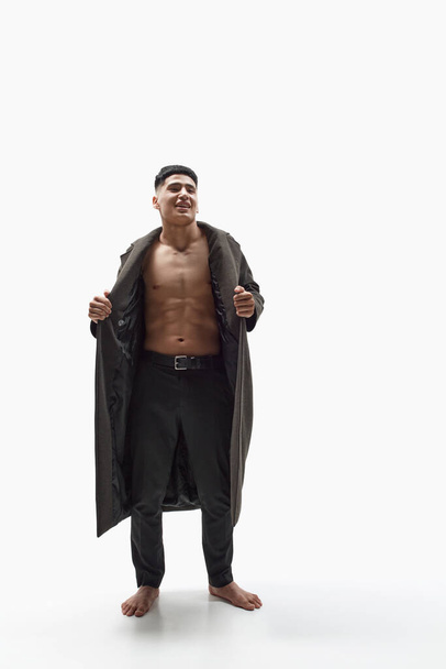Full-length portrait of smiling, handsome, young man posing shirtless with stylish coat and trousers against white studio background. Fashion, body aesthetics, beauty, mens health, emotions concept - Foto, immagini