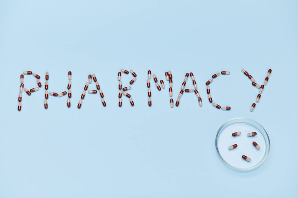 La parola FARMACIA disposta su fondo blu accanto alla capsula di Petri con capsule di gelatina medica con granuli terapeutici al loro interno. Ricevuto. Giornata mondiale del farmacista. Industria farmaceutica - Foto, immagini