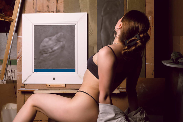 Sexy half naked girl is sitting in the dark attic of an art studio among forgotten paintings, arts and equipment. - Foto, imagen