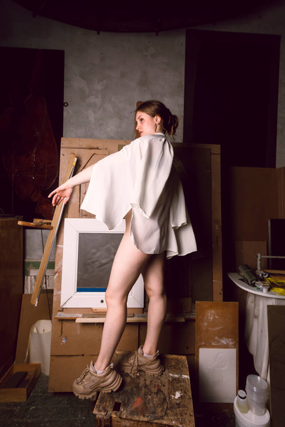 Sexy half naked girl is posing in the dark attic of an art studio among forgotten paintings, arts and equipment. - Photo, Image
