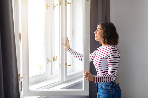 Beautiful Young Middle Eastern Female Opening Window In Room At Home, Smiling Arab Woman Breathing Fresh Air, Delighted Lady Standing With Eyes Closed, Enjoying Start Of New Day, Copy Space - Photo, Image