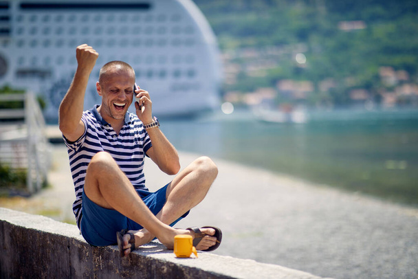 A young man is talking on the smartphone while taking a rest on the quay on a beautiful sunny day during a vacation on the seaside. Vacation, seaside, tourists - Photo, Image