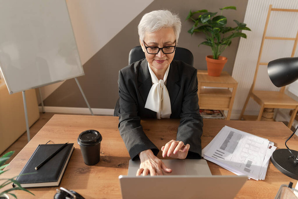 Confident stylish european middle aged senior woman using laptop at workplace. Stylish older mature 60s gray haired lady businesswoman sitting at office table. Boss leader teacher professional worker - 写真・画像