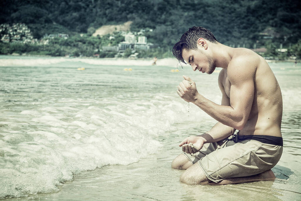 Handsome young man sitting on a beach playing with sand in Phuket Island, Thailand, shirtless wearing boxer shorts, showing muscular fit body - Foto, afbeelding