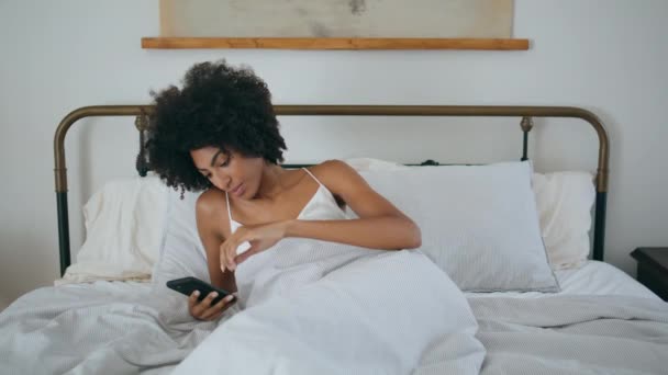 Прокручивание телефона леди в спальне. Спокойная курчавая брюнетка проверяет социальные сети, используя смартфон в постели. Расслабляющая женщина, занимающаяся серфингом в интернете, разговаривает по телефону дома. Сексуальная африканская девушка отдыхает подушку - Кадры, видео