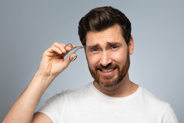 Hair removal and self care concept. Handsome bearded middle aged man tweezing eyebrows, standing over grey background, studio shot. - Foto, Bild
