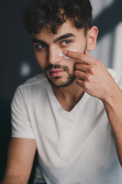 Közelkép a fiatal kaukázusi férfi néz el alkalmazása hidratáló arc ránctalanító krém az egészséges puha bőr. Bőrápolás, szépségápolás. Arcbőrápolás - Fotó, kép