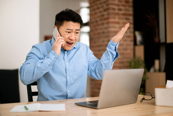 Emotional angry asian middle aged businessman working from home, having phone conversation, looking at laptop screen and gesturing, copy space - Foto, Bild