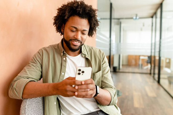 Smiling african american business man with smartphone in hand looking screen and sitting indoor, male office employee, entrepreneur using smartphone, chatting and texting online - Foto, Imagem
