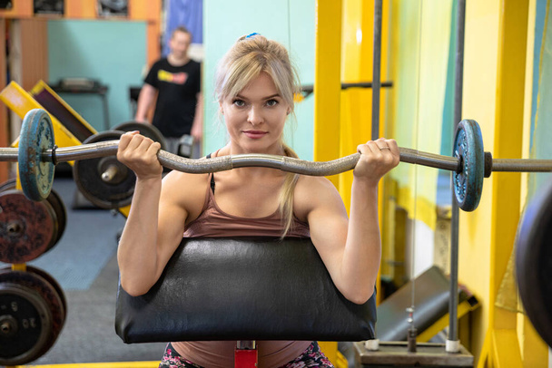 A girl at the gym holds a barbell in front of her. The woman raises the barbell to chest height. A mirror can be seen in the background. In the mirror is the reflection of a blurry person in a black t - Foto, Bild