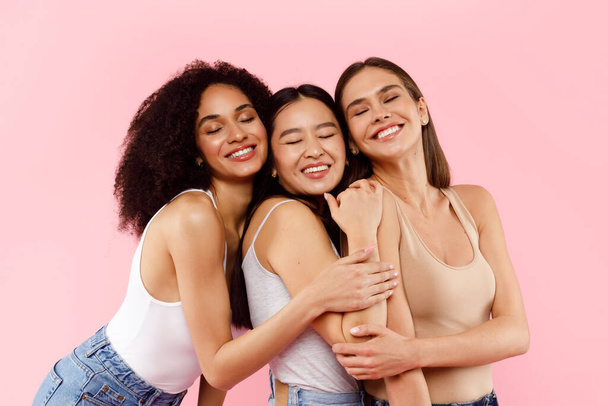 Three happy multiracial female best friends, embracing and smiling with closed eyes, posing in casual, standing in studio over pink background - Fotografie, Obrázek