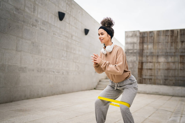 Eine erwachsene kaukasische Athletin trainiert mit elastischen Gummiwiderstandsbändern im Freien in der Stadt auf Betonhintergrund, der sich in fröhlichem brünetten Gesundheits- und Fitnesskonzept ausdehnt. - Foto, Bild