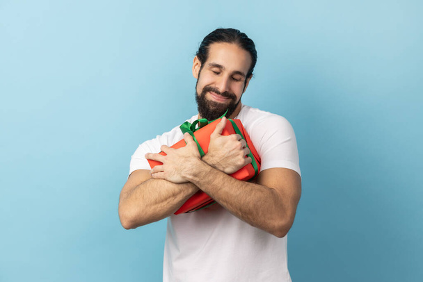 Long-awaited present. Portrait of delighted handsome man with beard wearing white T-shirt embracing red gift box, satisfied with surprise. Indoor studio shot isolated on blue background. - Zdjęcie, obraz