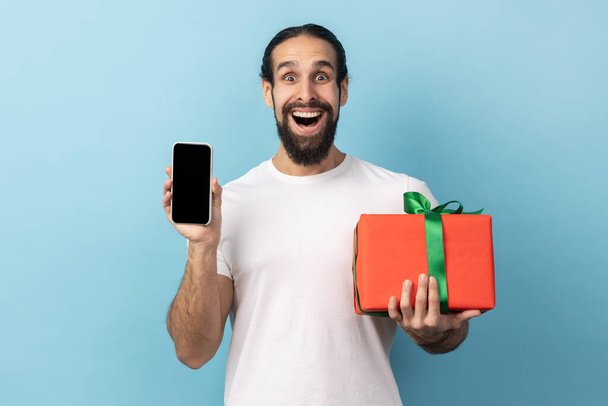 Unbelievable present, bonus for mobile user. Man with beard holding gift box and cell phone with mock up, blank display for advertise. Indoor studio shot isolated on blue background. - 写真・画像