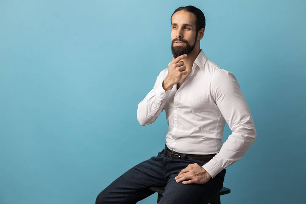 Portrait of man with beard wearing white shirt and trousers sitting with pensive thoughtful facial expression, looking away and holding chin. Indoor studio shot isolated on blue background. - Foto, Bild