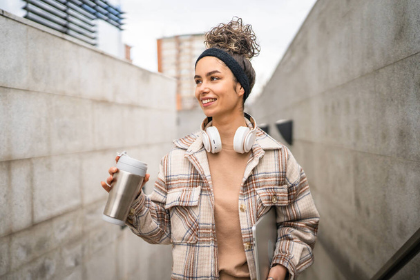 Una mujer joven adulta estudiante de pie o caminar al aire libre sostener ordenador portátil y termo taza de café persona real copiar espacio sonrisa feliz confiado con auriculares en el día - Foto, imagen