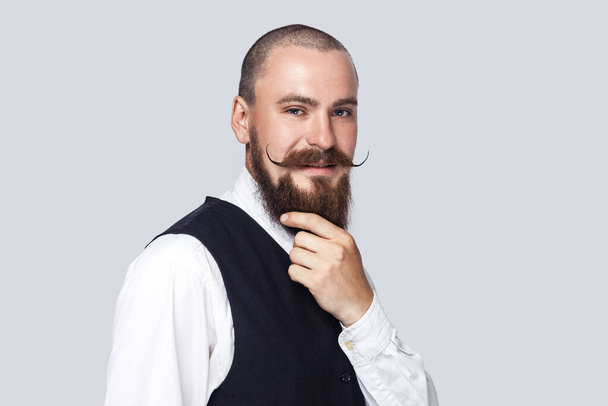 Portrait of smiling happy handsome bearded man with mustache looking at camera with pleasant facial expression, touching his beard. Indoor studio shot isolated on gray background. - Фото, изображение