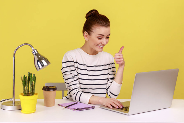 Happy woman office manager sitting office workplace, showing thumbs up, satisfied with excellent business result, having video call. Indoor studio studio shot isolated on yellow background. - 写真・画像