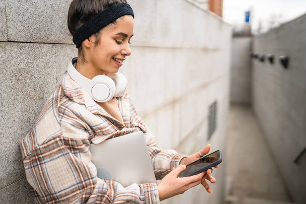 Una mujer joven adulta estudiante de pie o caminar al aire libre ordenador portátil uso teléfono inteligente para mensajes de texto SMS en línea persona real copiar espacio sonrisa feliz confiado con auriculares en el día - Foto, imagen