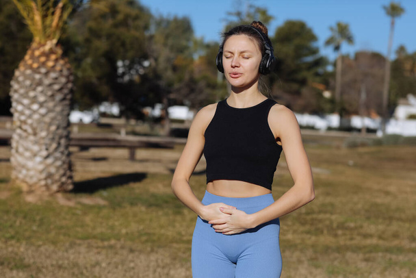 Concentrated young woman wearing headphones doing yoga meditation with online tutorial outdoors at park with palm trees. Listening music, harmony, calming, spirituality concept. World health day. - Valokuva, kuva