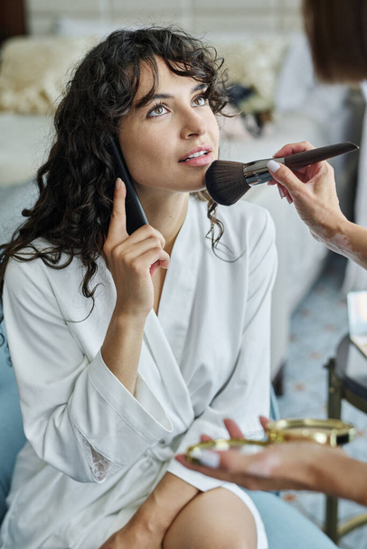 Mujer joven con el pelo ondulado largo oscuro hablando en el teléfono inteligente y mirando artista de maquillaje aplicando polvo suelto en su cara con cepillo - Foto, Imagen