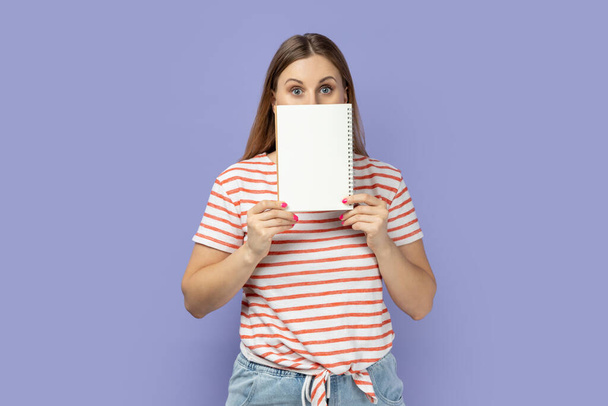 Portrait of shocked shy blond woman wearing striped T-shirt holding organizer with empty paper with advertising area, looking at camera. Indoor studio shot isolated on purple background. - Photo, Image