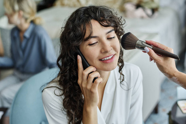 Happy young bride with dark long wavy hair speaking on smartphone while makeup artist with brush applying powder on her cheek - Foto, immagini
