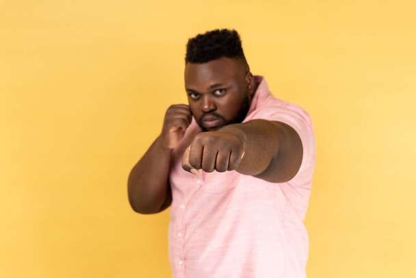 I'll hit you. Portrait of bearded man wearing pink shirt standing with raised fists boxing gesture, ready to punch, ready to struggle, fighting spirit. Indoor studio shot isolated on yellow background - Фото, изображение