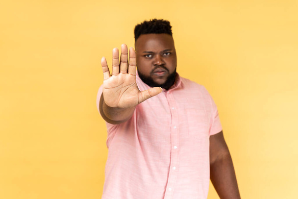 Portrait of dissatisfied man wearing pink shirt standing with disgust expression, frowning showing stop gesture with hand. Indoor studio shot isolated on yellow background. - Foto, immagini