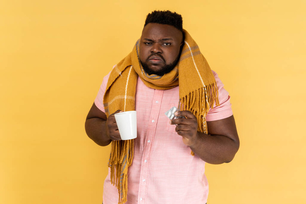 Portrait of sick unhealthy bearded man wearing pink shirt wrapped in scarf holding tea and pills, feeling despair about influenza. Indoor studio shot isolated on yellow background. - Foto, imagen