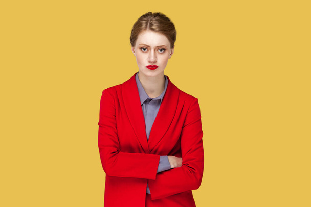 Portrait of serious strict woman with red lips standing standing with crossed arms, looking at camera, having bossy expression, wearing red jacket. Indoor studio shot isolated on yellow background. - Photo, image