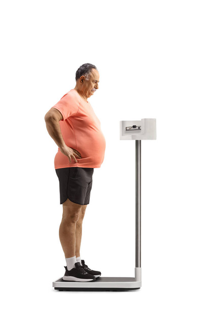 Worried mature man standing on a medical weight scale isolated on white background - 写真・画像