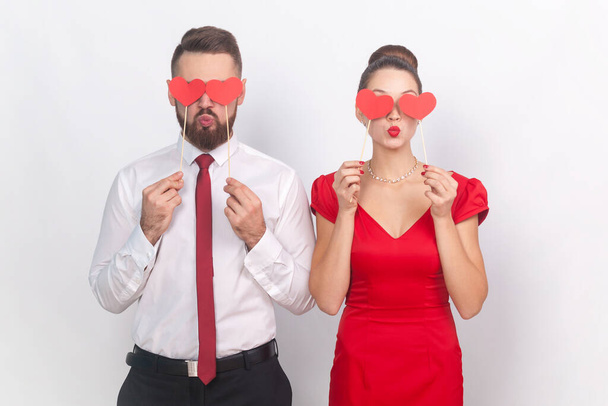 Flirting man in white shirt and woman in red dress standing together, holding little hearts, covering eye with love symbols on stick, sending air kisses. Indoor studio shot isolated on gray background - Photo, Image
