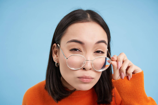 Gros plan de la femme asiatique dans des lunettes, pensant, regardant la caméra avec expression réfléchie du visage, debout sur fond bleu. - Photo, image
