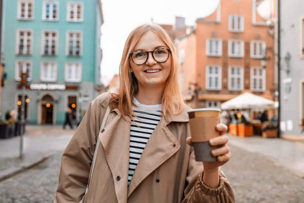 Молодая улыбающаяся женщина в пальто с чашкой кофе на вечерней городской улице в старом городе Гданьска. Путешествует по Европе осенью. Высокое качество фото - Фото, изображение
