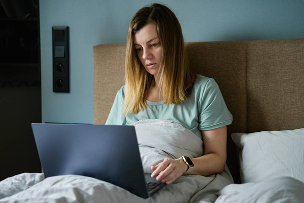 Millennial female freelancer lying in bed and using laptop. Woman work from home. Comfortable workplace. - Fotoğraf, Görsel