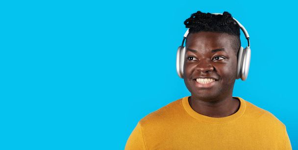 Portrait Of Smiling Black Young Man Listening Music In Wireless Headphones, Happy Cheerful African American Guy Enjoying Favorite Songs And Looking Away While Posing Over Blue Background, Panorama - Zdjęcie, obraz