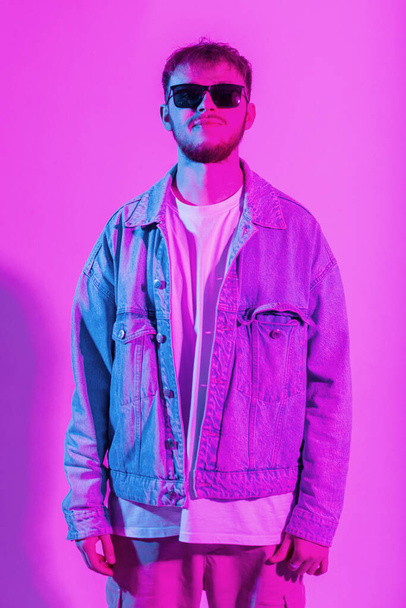Модный креативный молодой красивый мужчина в темных очках в джинсовой модной куртке в красочной студии с розовым и голубым светом - Фото, изображение